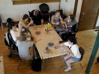 Top view of a large  square wooden  table surrounded by three people using wheelchairs painting with four people without disabilities