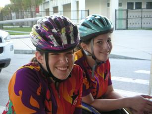 Two girls who use wheelchairs are dressed in colorful cycling shirts and helmets as they smile at the camera.