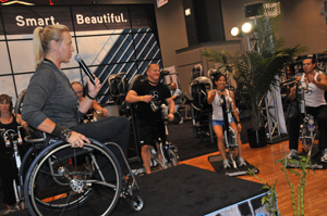 A women in a wheelchair is leading a group of people through an exercise class on an upper body cycle.